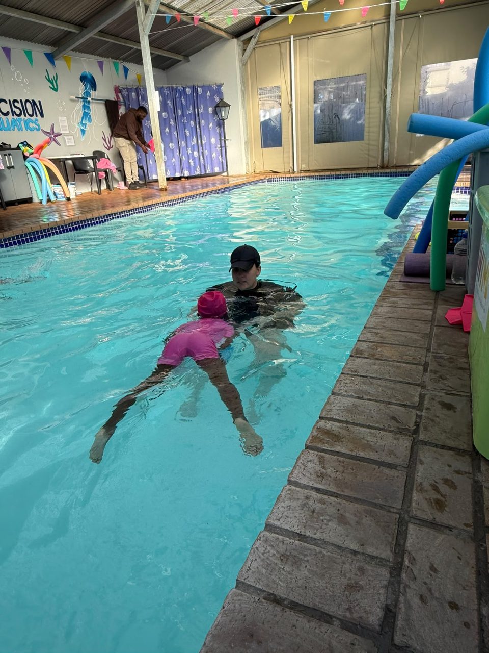 Instructor teaching a child to swim at Precision Aquatics' indoor pool, with the Precision Aquatics wall signage visible